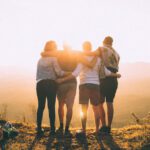 Travel Challenge - four person hands wrap around shoulders while looking at sunset